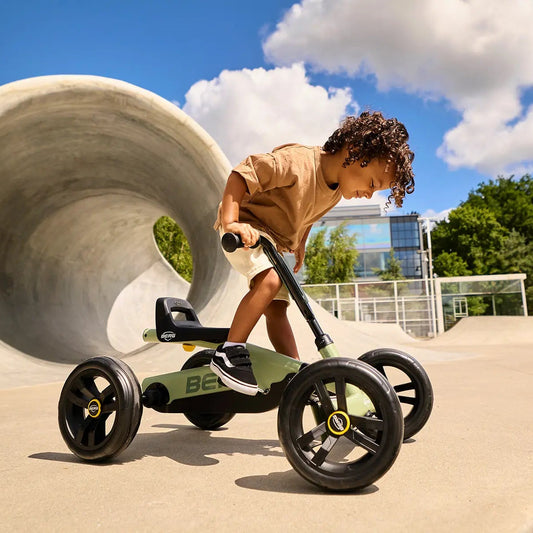 A child riding a green and black three-wheel tricycle in an outdoor concrete skatepark under a blue sky with clouds.