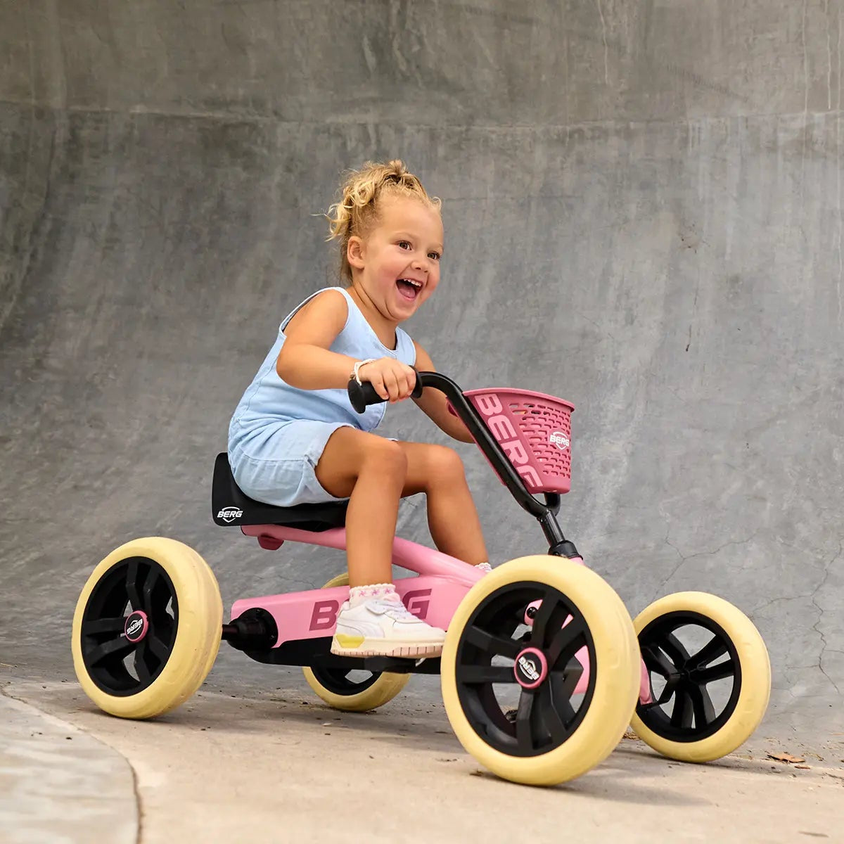A young child riding a pink and black tricycle with yellow wheels in front of a concrete wall.