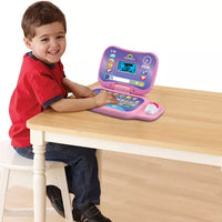 toddler sitting at table with pink and purple toy laptop 