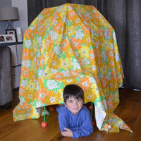 Child sitting inside a colorful floral tent on a wooden floor.