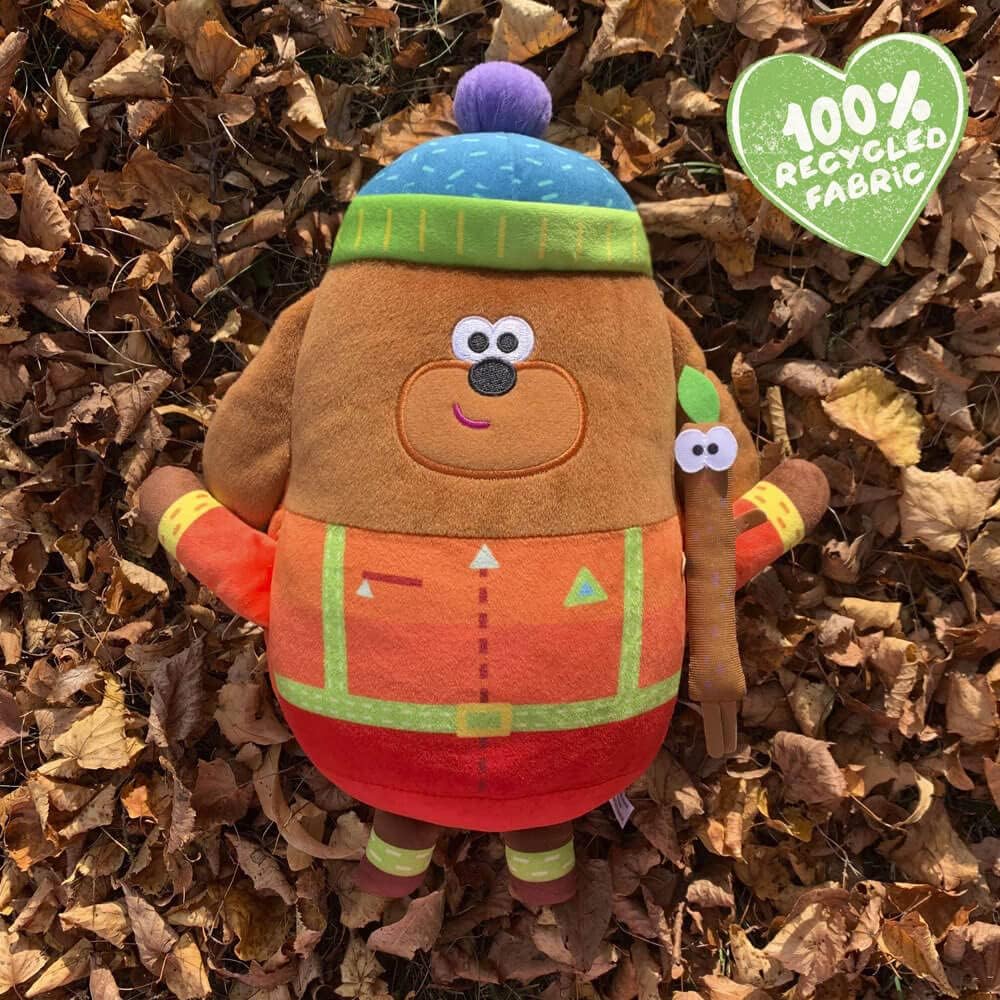 Plush toy bear wearing a colorful hat and scarf on a bed of leaves with a '100% Recycled Fabric' label.