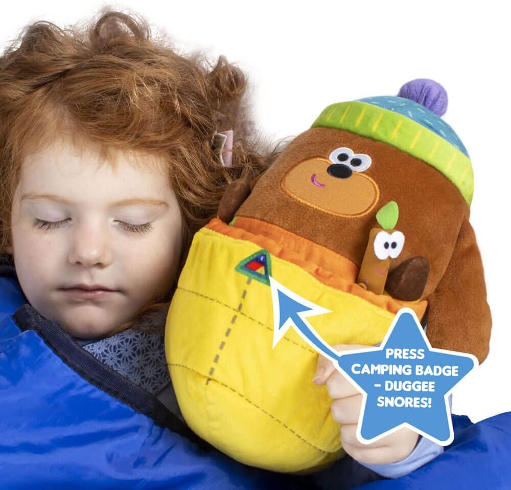 Child sleeping with a plush toy resembling Duggee from Ben & Holly's Little Kingdom, with a blue star label indicating a feature.
